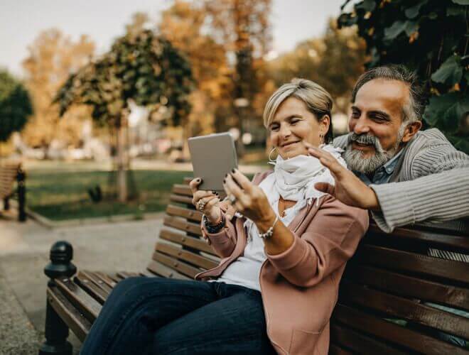 Middle aged couple sitting on a park bench having a video call on a tablet