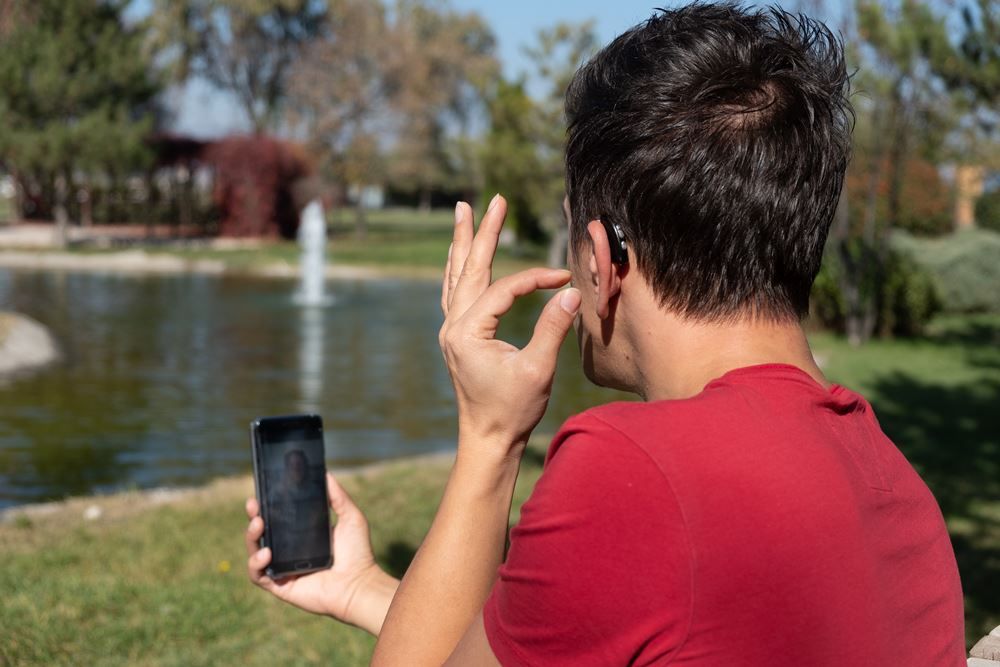 Young man adjusting his hearing aid with his phone by a pond